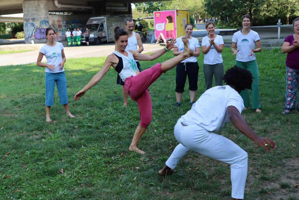Auf dem Foto sind mehrere Personen auf einer Wiese zu sehen. Im Vordergrund macht eine Frau in Sport-Top und linker Sporthose gerade einen Kick, ein Mann in weißem Capoeira-Outfit weicht aus. Im Hintergrund klatschen und lachen mehrere Personen.