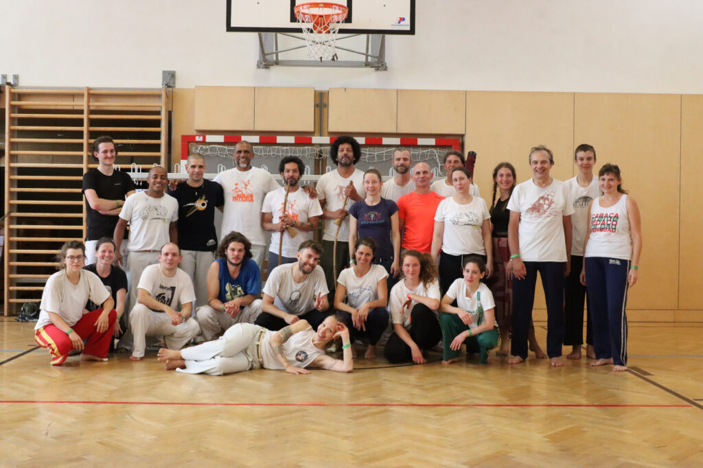 Eine Gruppe von rund 25 Menschen in Sportkleidung posiert in einem Turnsaal für ein Gruppenfoto. Zwei Männer halten jeweils ein Berimbau, ein brasilianisches Musikinstrument, in der Hand.