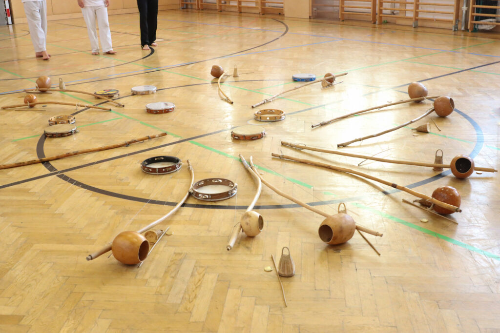 Auf dem Boden eines Turnsaals liegen viele Instrumente im Kreis: Pandeiros (eine Art von Tamburin) und Berimbaus (ein brasilianisches Instrument, das in der Capoeira verwendet wird).